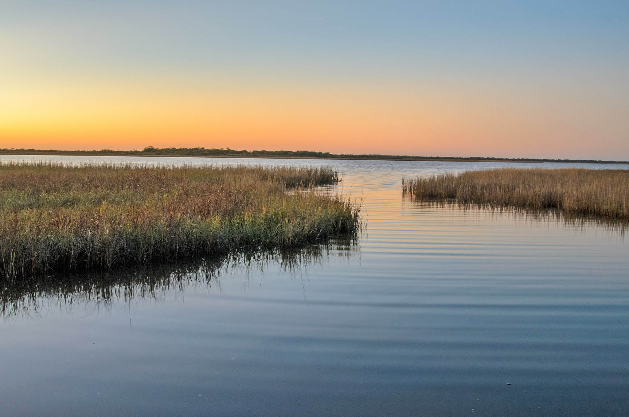 An expanse of dense coastal wetlands intermixed with open water along the Texas Gulf Coast at sunset; the sky is orange.