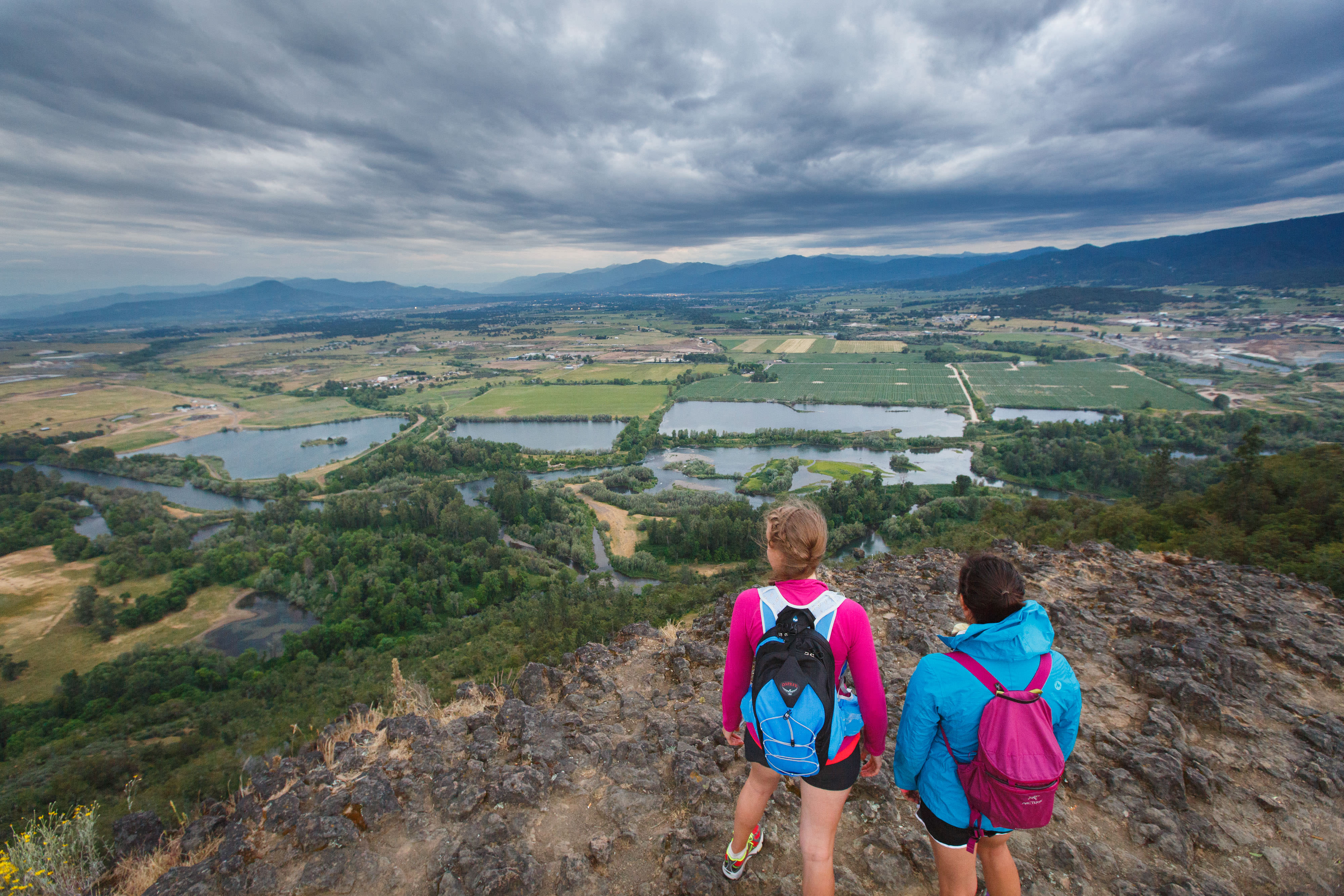 Two people standing on a rock outcrop looking out over forests and farms.