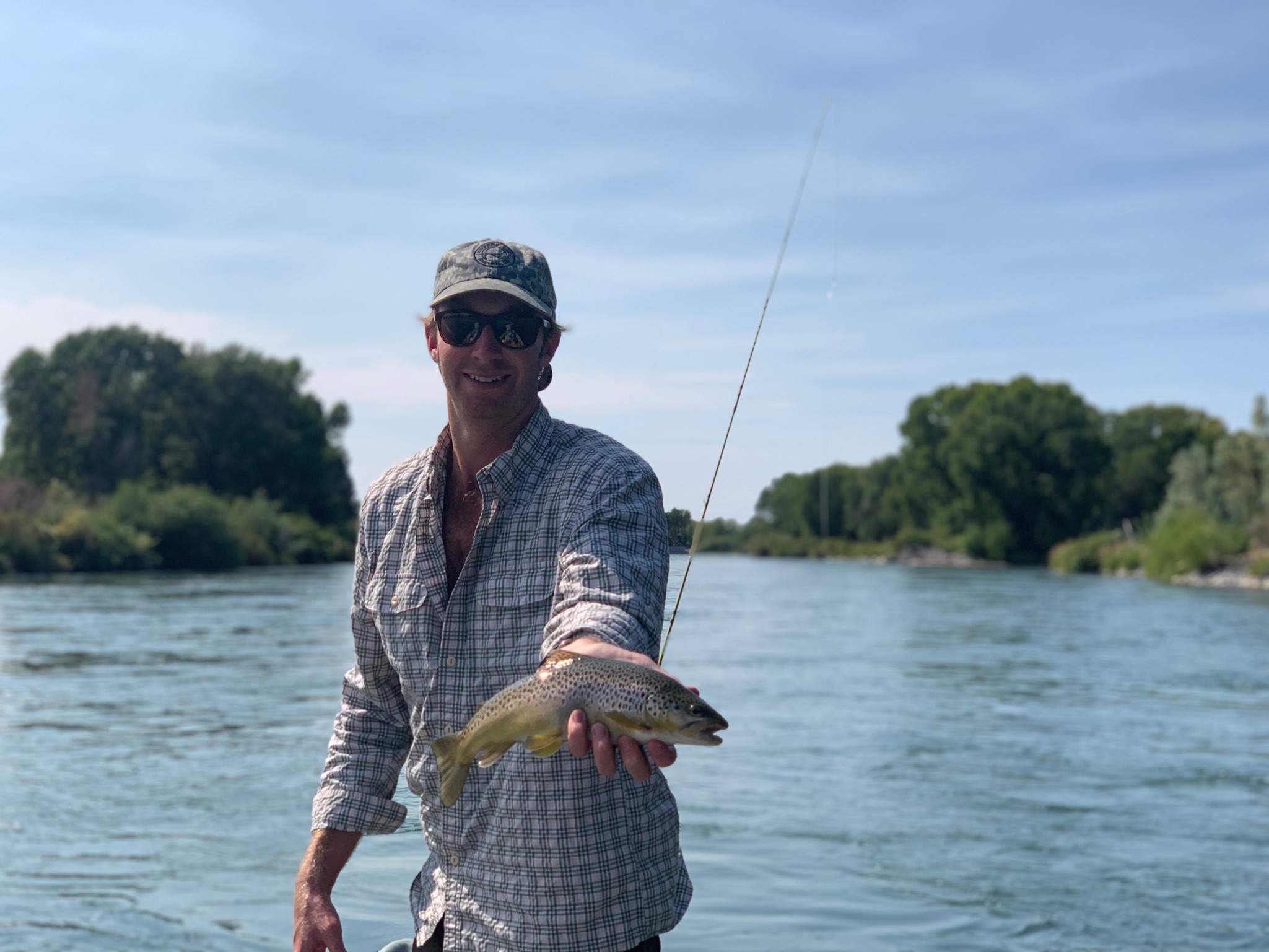 Thomas Reddick holding out a fish with one hand, stands in front of a water body with a fishing pole  behind him.