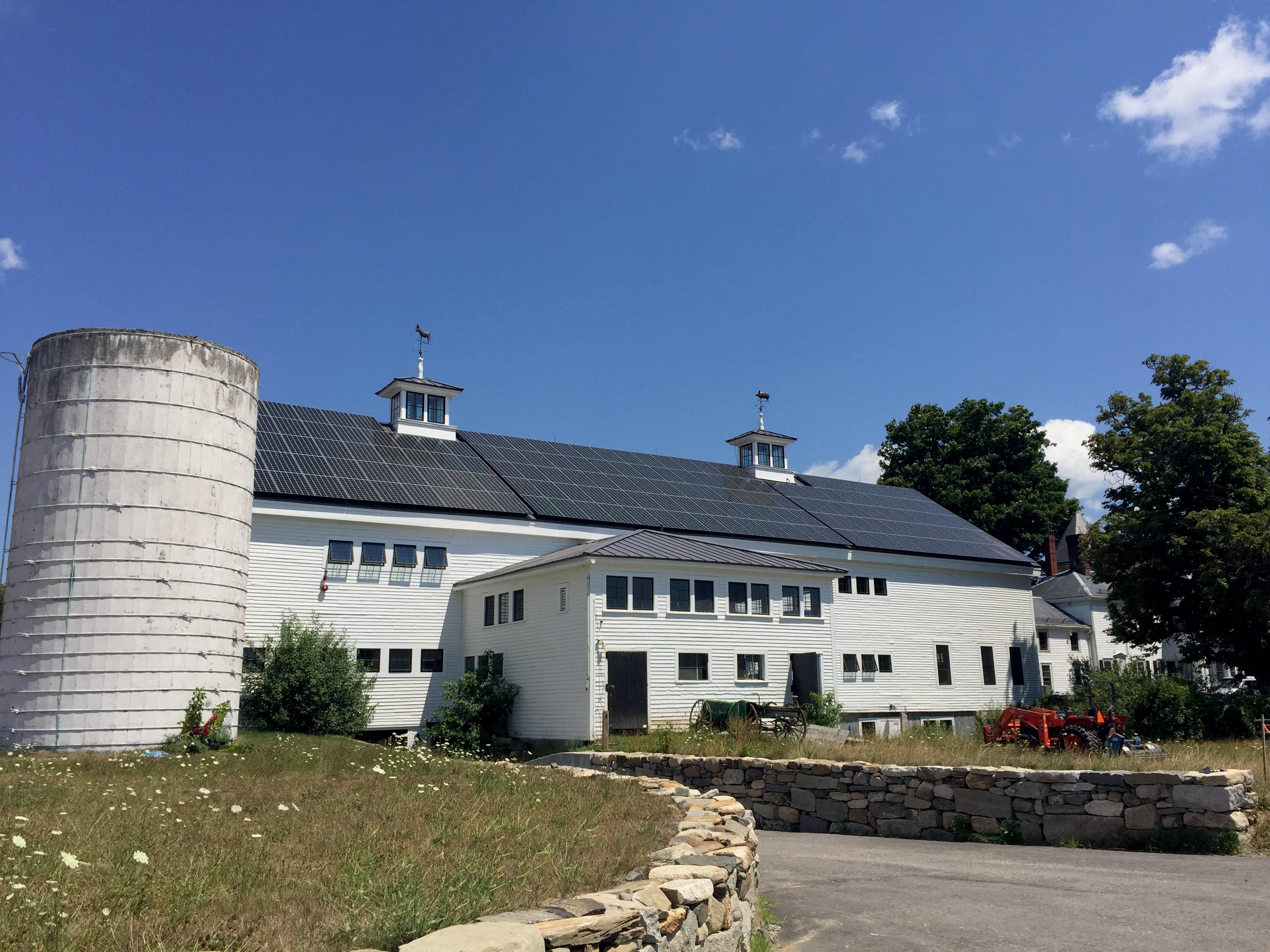 A large white barn with a roof covered in solar panels. A white silo stands to the left and a stonewall to the right.