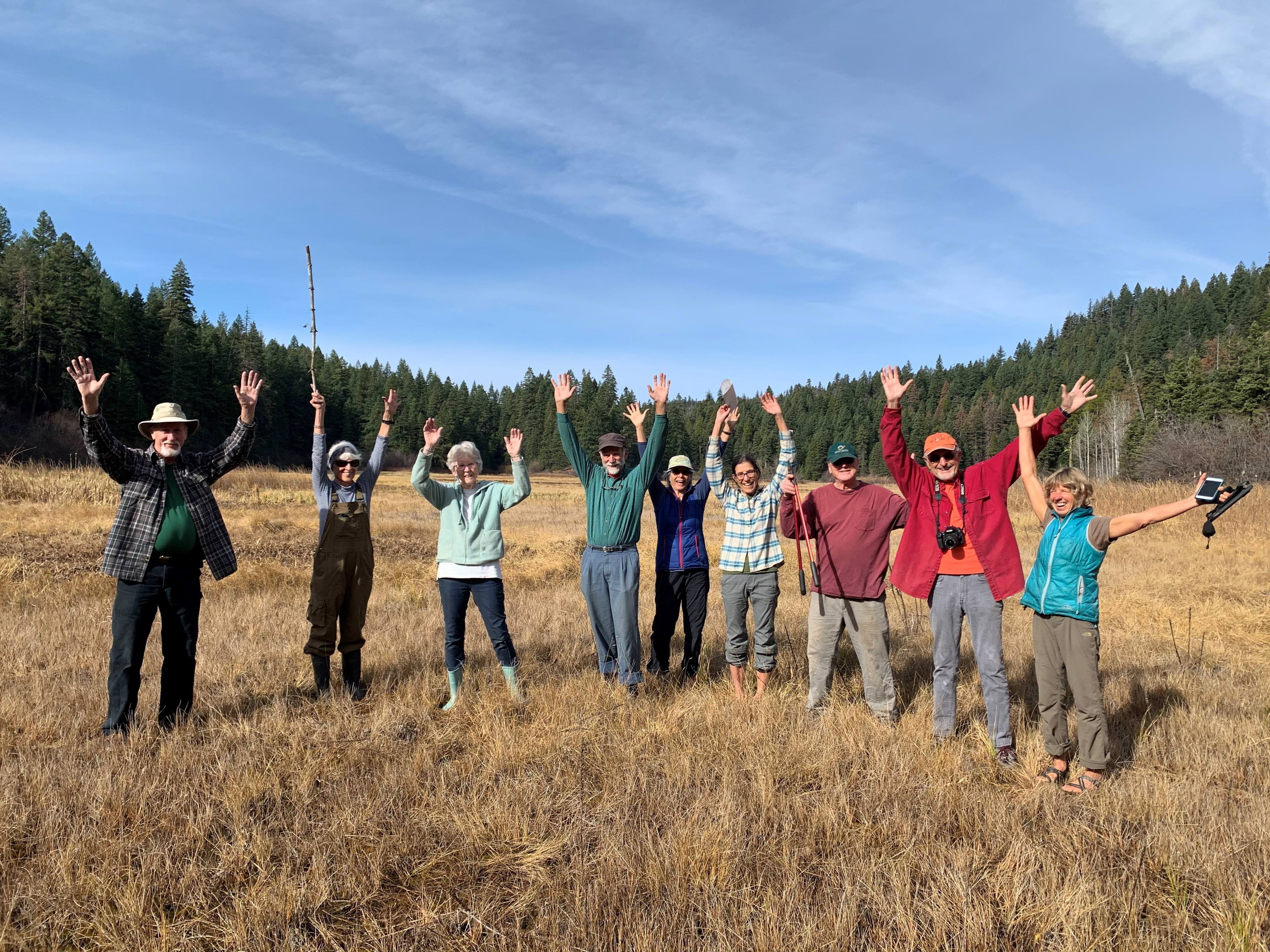 Nine people standing in a grassy field in colorful hiking clothing with their hands in the air, forest in the background.