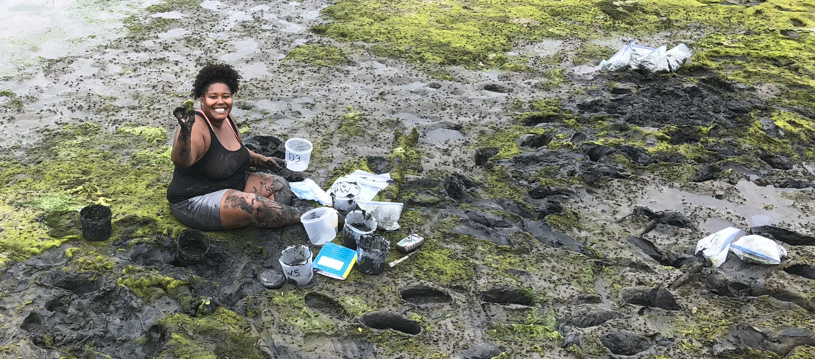 Tiara Moore smiles and holds up a plant sample as she sits in a muddy area with deep footprints and green growth