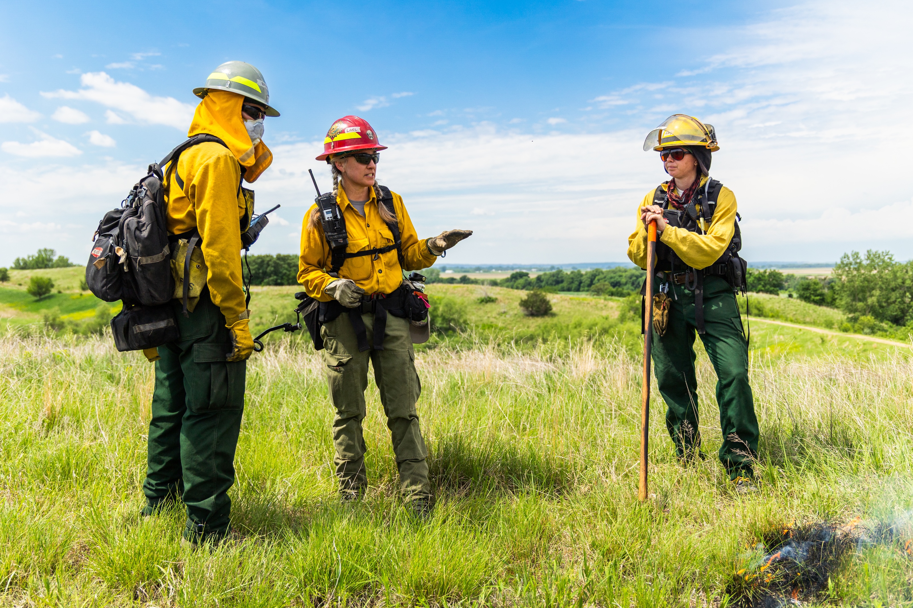 Three people dressed in yellow and wearing hardhats meet in a green, grassy field.
