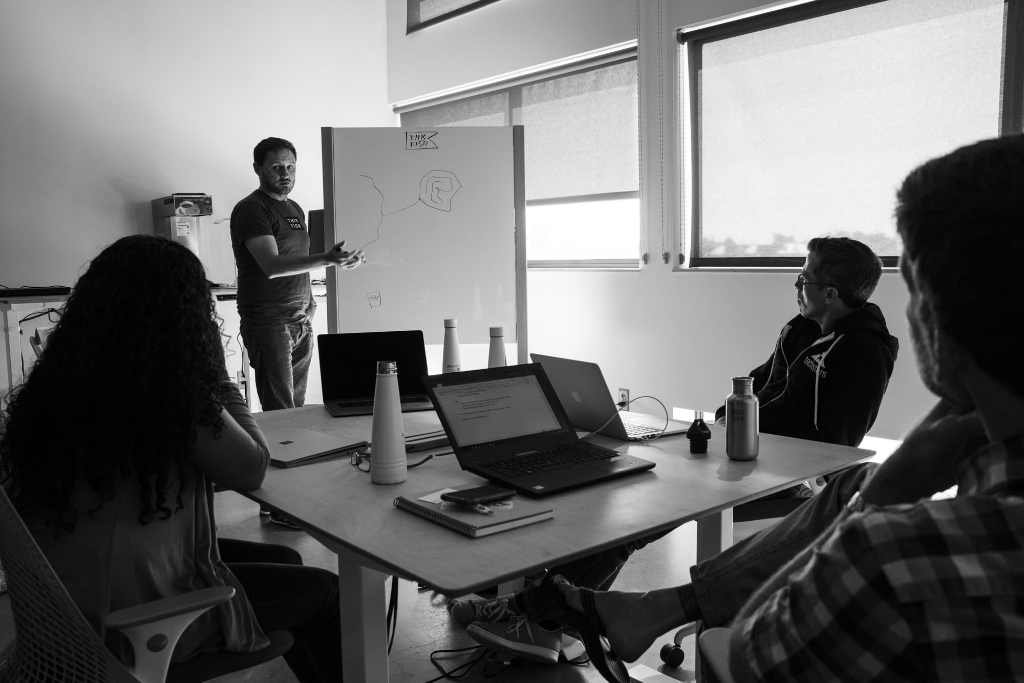 A man stand next to a whiteboard, gesturing toward the simple drawing on it, while others look on