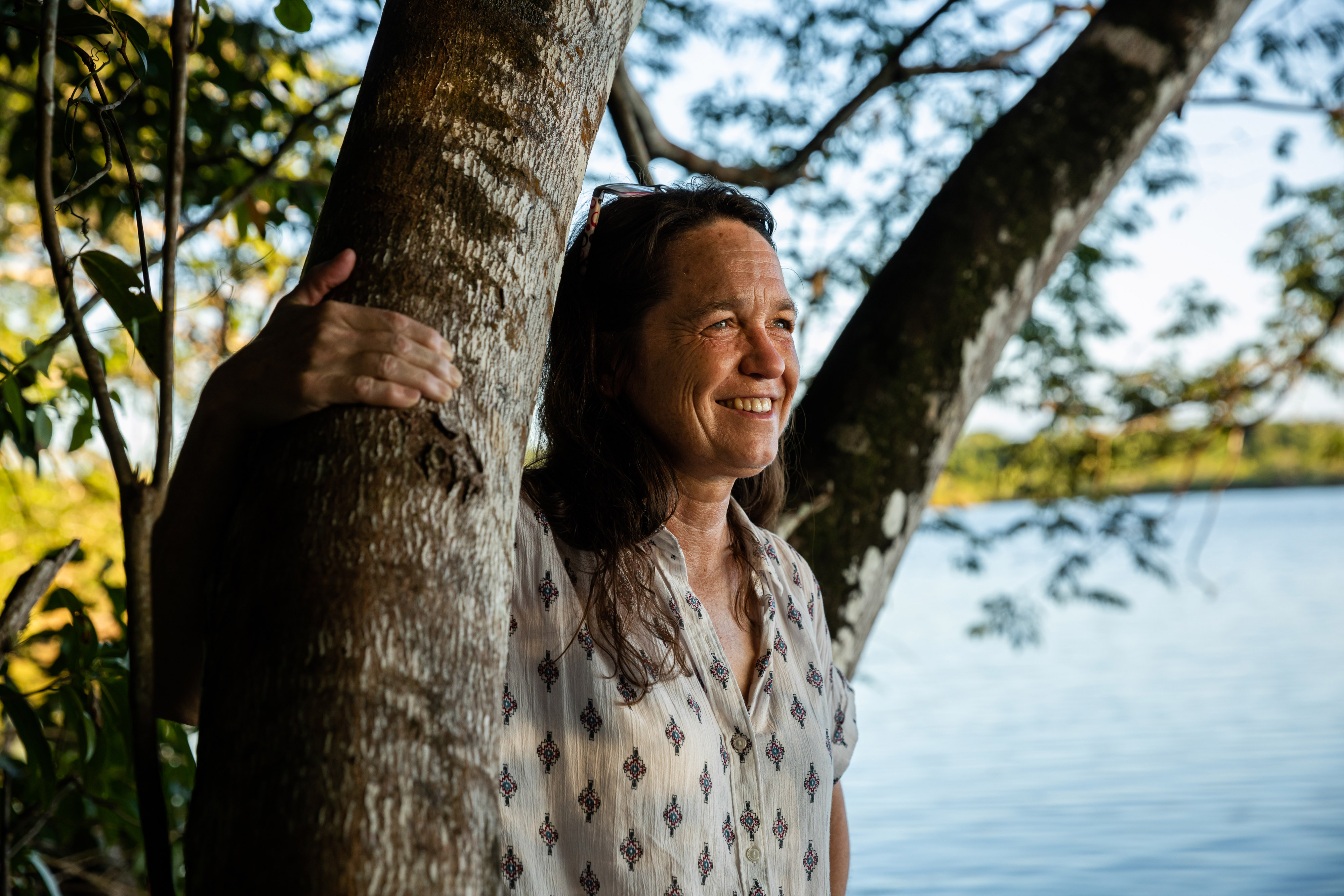 A woman smiles as she stands near a tree and a blue pool of water.