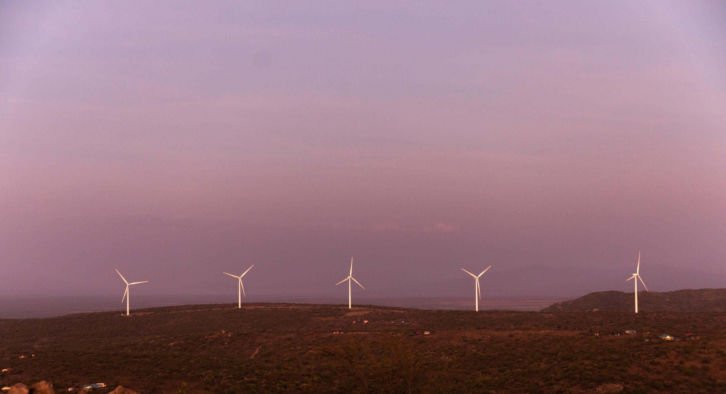 A row of wind turbines stand along a ridge under a pink sky.