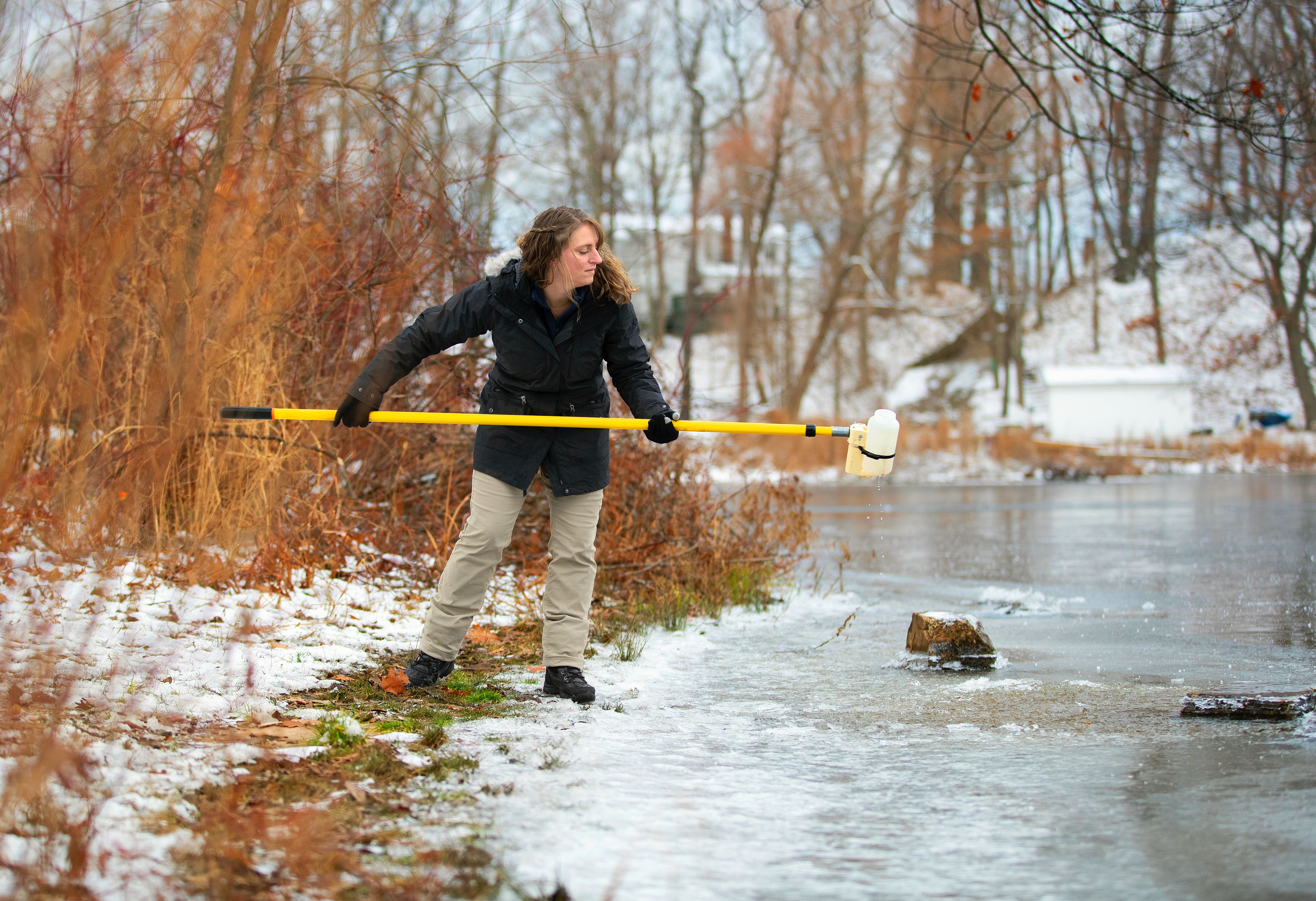 Brittney Rogers stands on the bank of the Salmon River on the lefthand side, using a yellow pole and bottle attached to it to collect a water sample. 