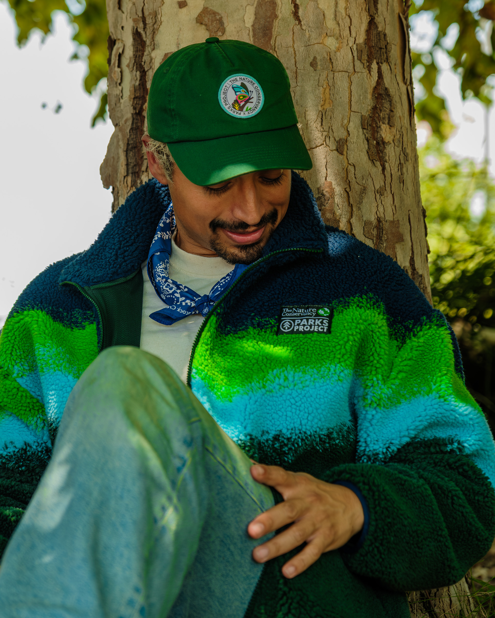 A man sits against the trunk of a birch tree looking at the Parks Project x TNC chest patch on his fleece jacket. His has short bleached hair and wears a green baseball style hat with the Parks Project x TNC 75th anniversary collab patch.