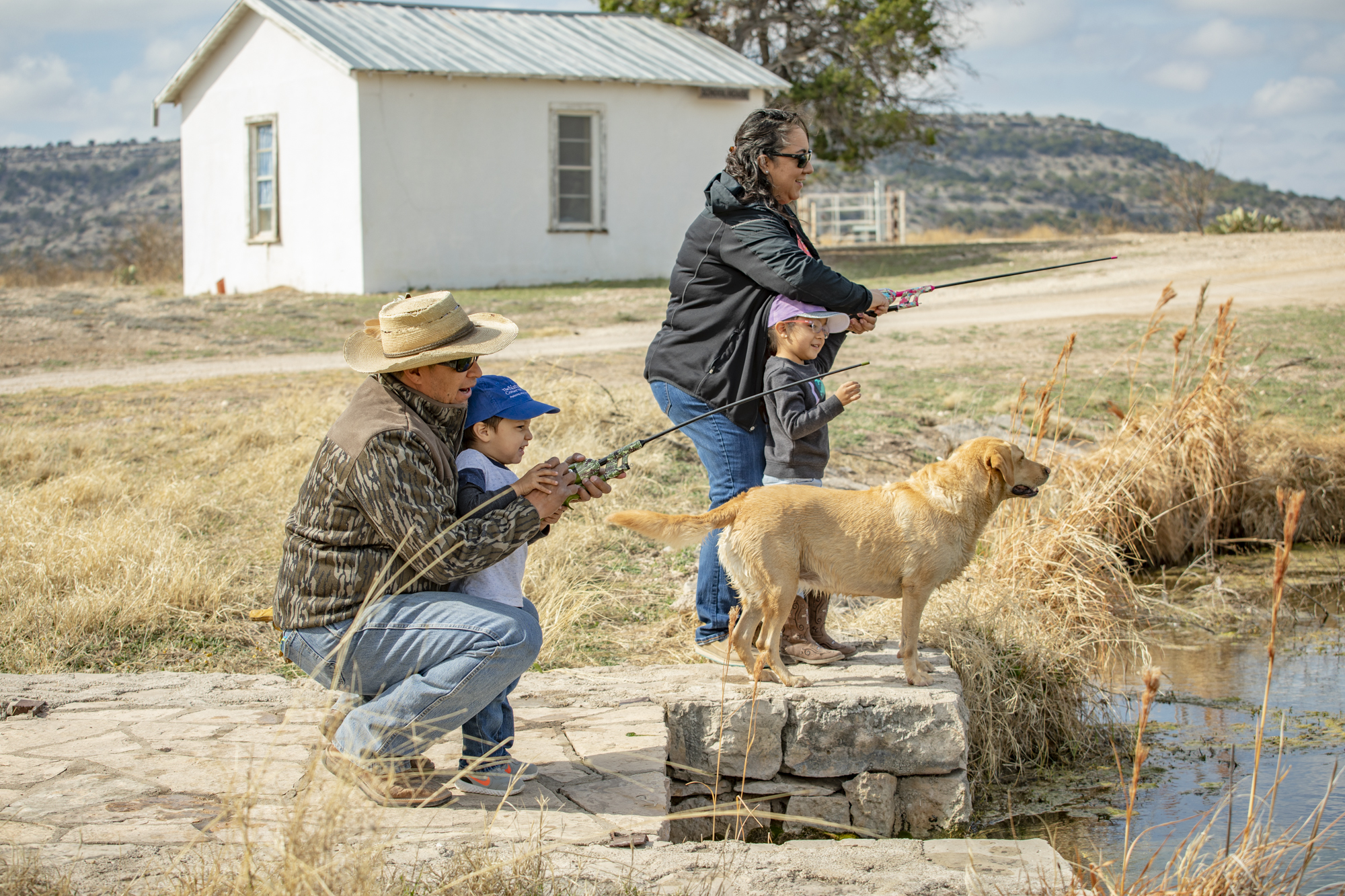 A man and women help a little boy and girl cast their rods to fish in a pond while their yellow labrador watches intently.