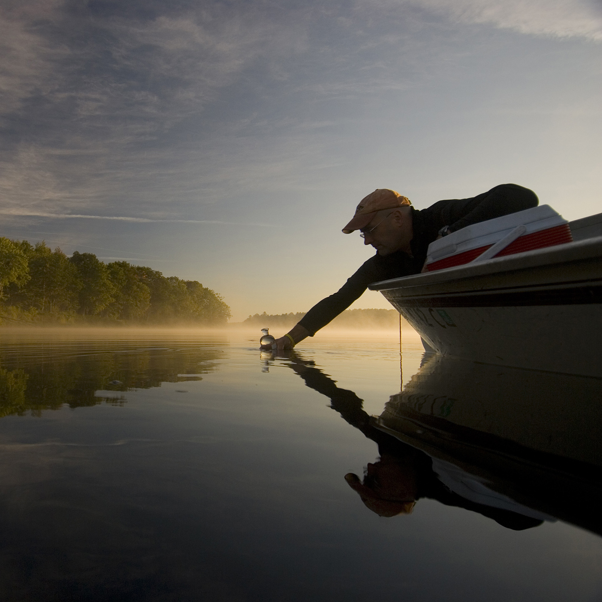 A man reaches out of a small boat and dips a glass bottle into the water.