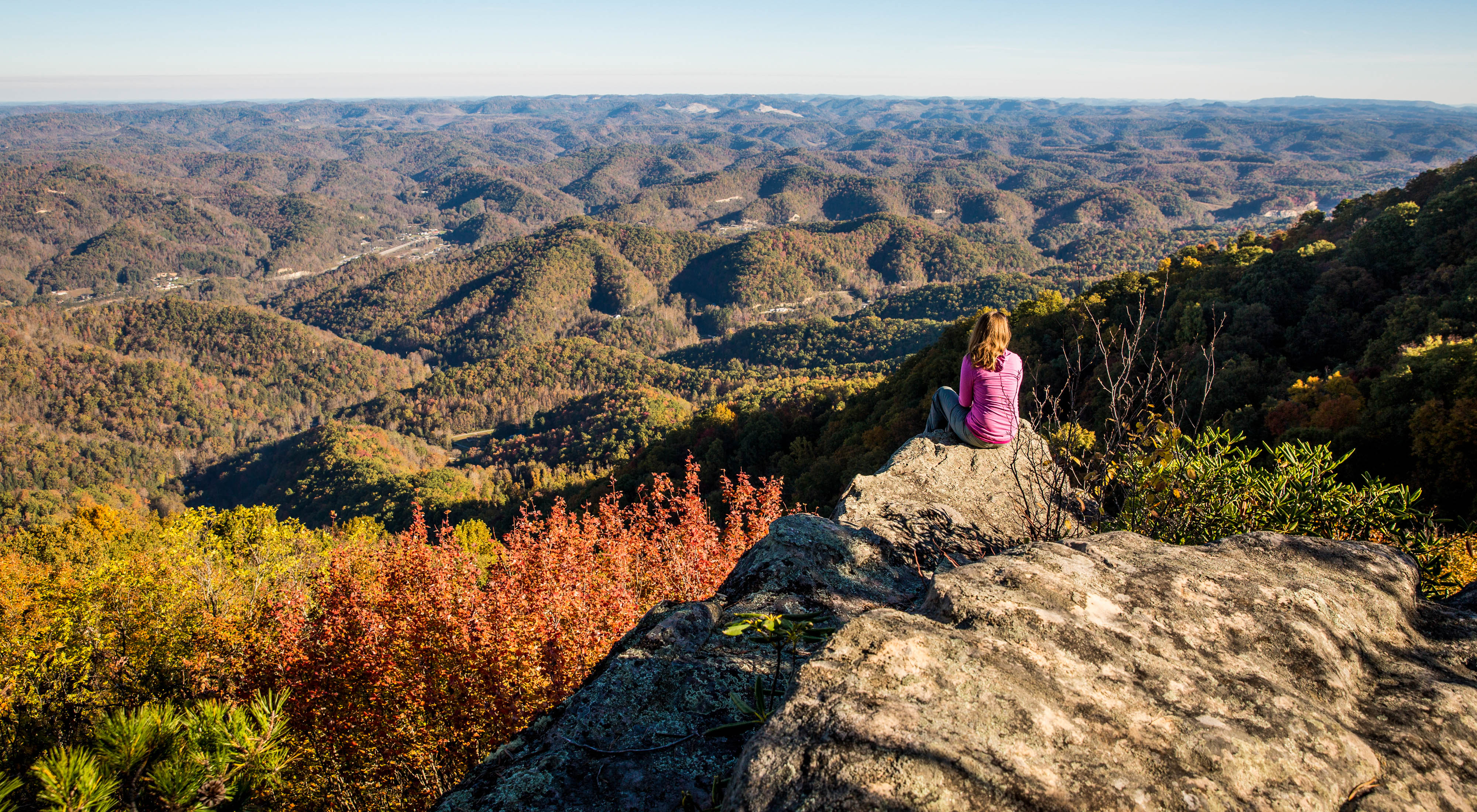 A woman sits on a rock outcropping and looks out across a vast mountainous landscape.