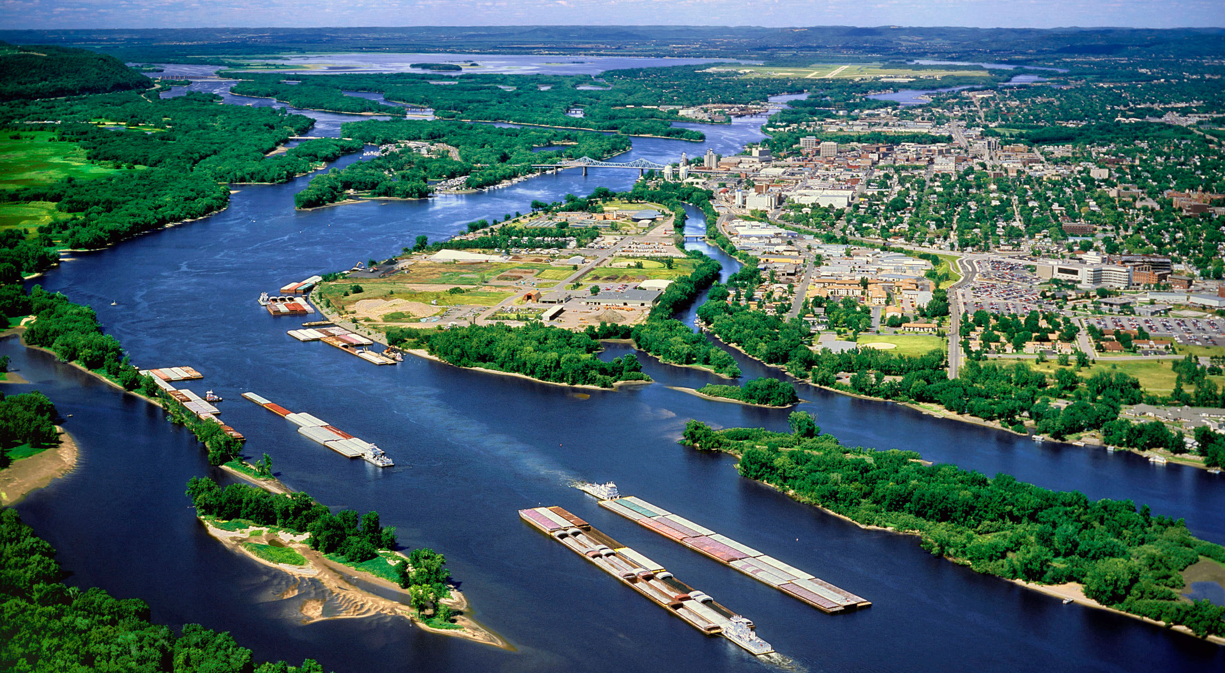 An aerial view of barges on the Mississippi river as it curves around populated and forested areas.