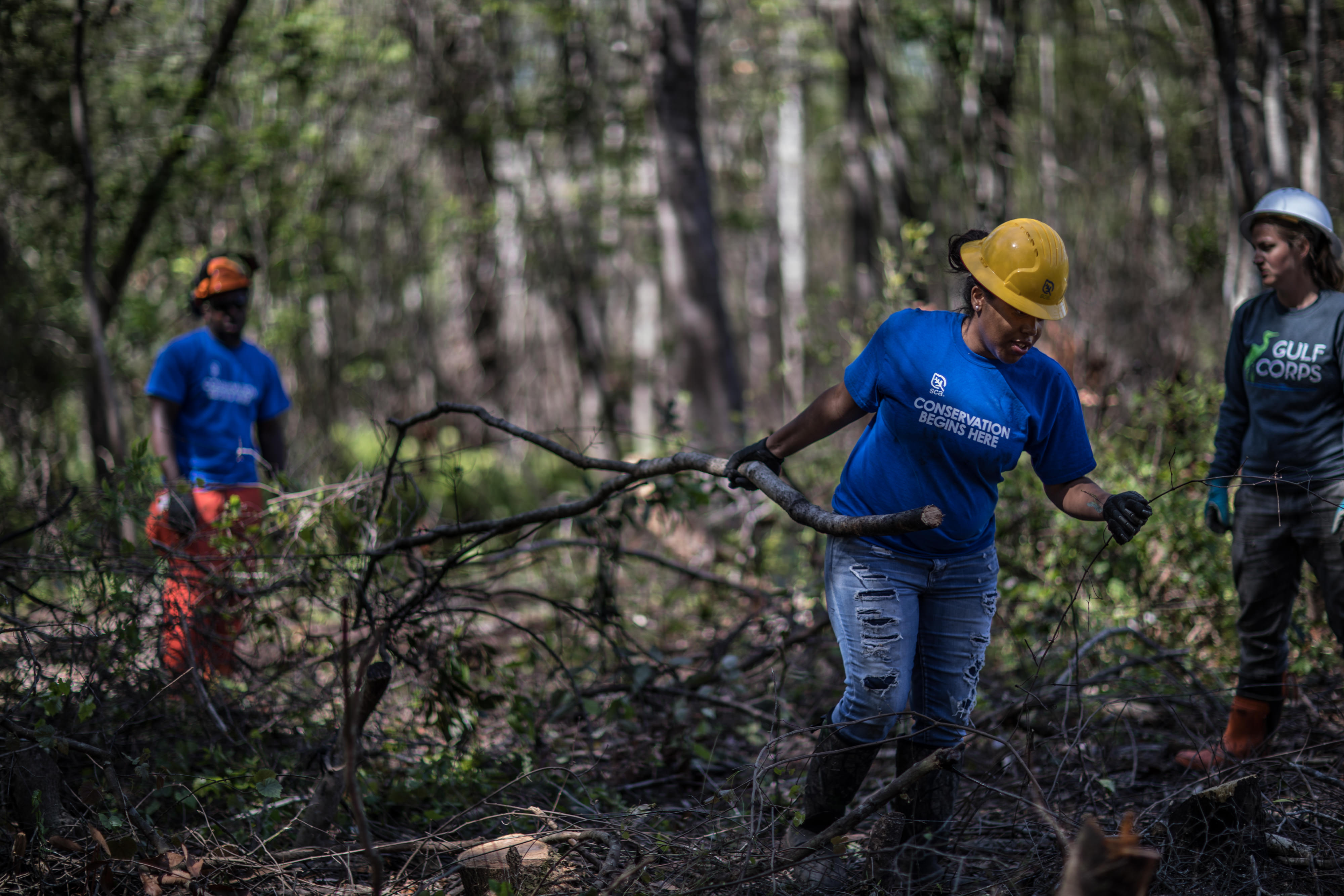 Three young people in hard hats, gloves and conservation corps shirts, clearing branches in a forest.