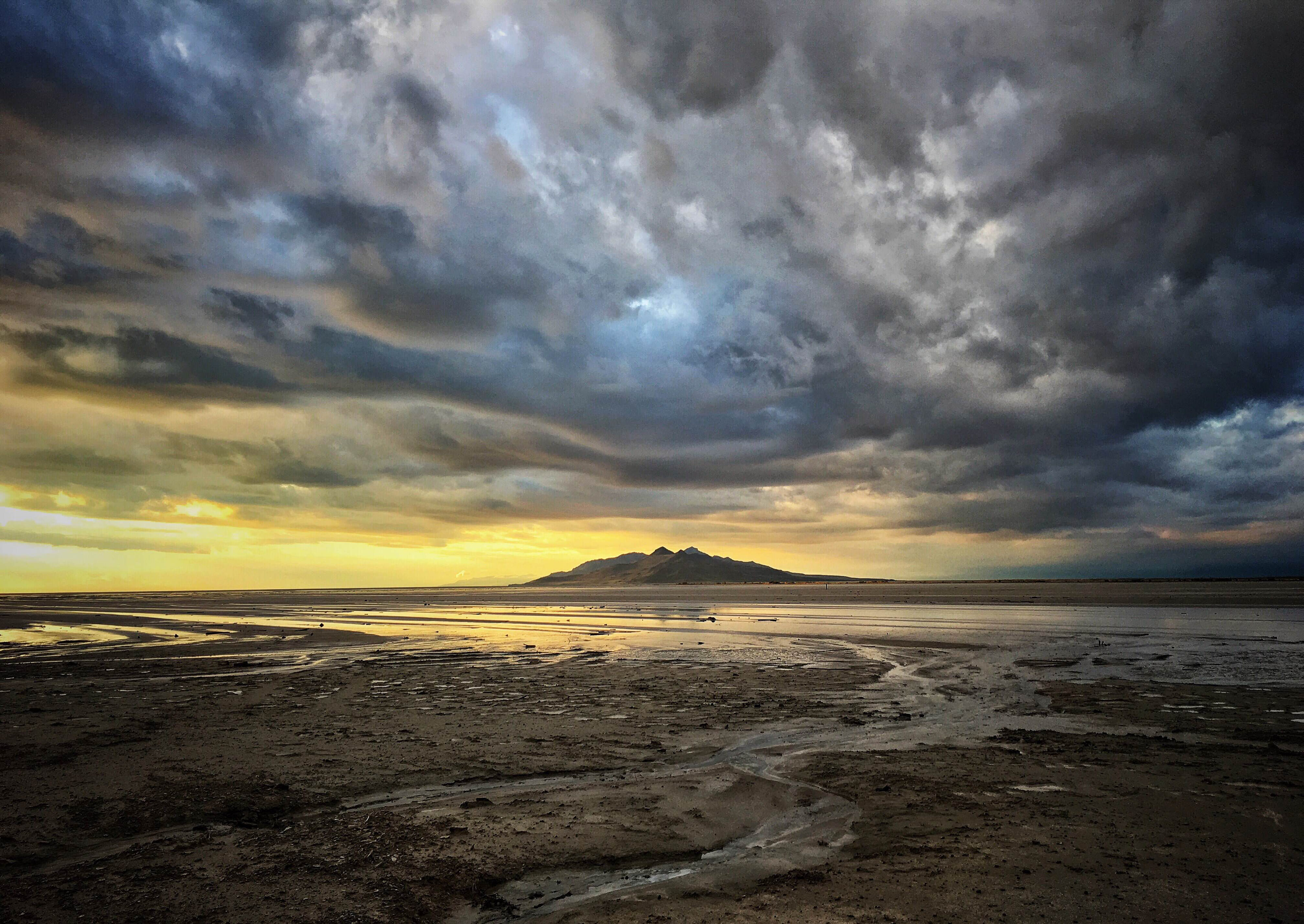Shallow water in the great salt lake under dramatic cloudy skies with a hill in the distance.