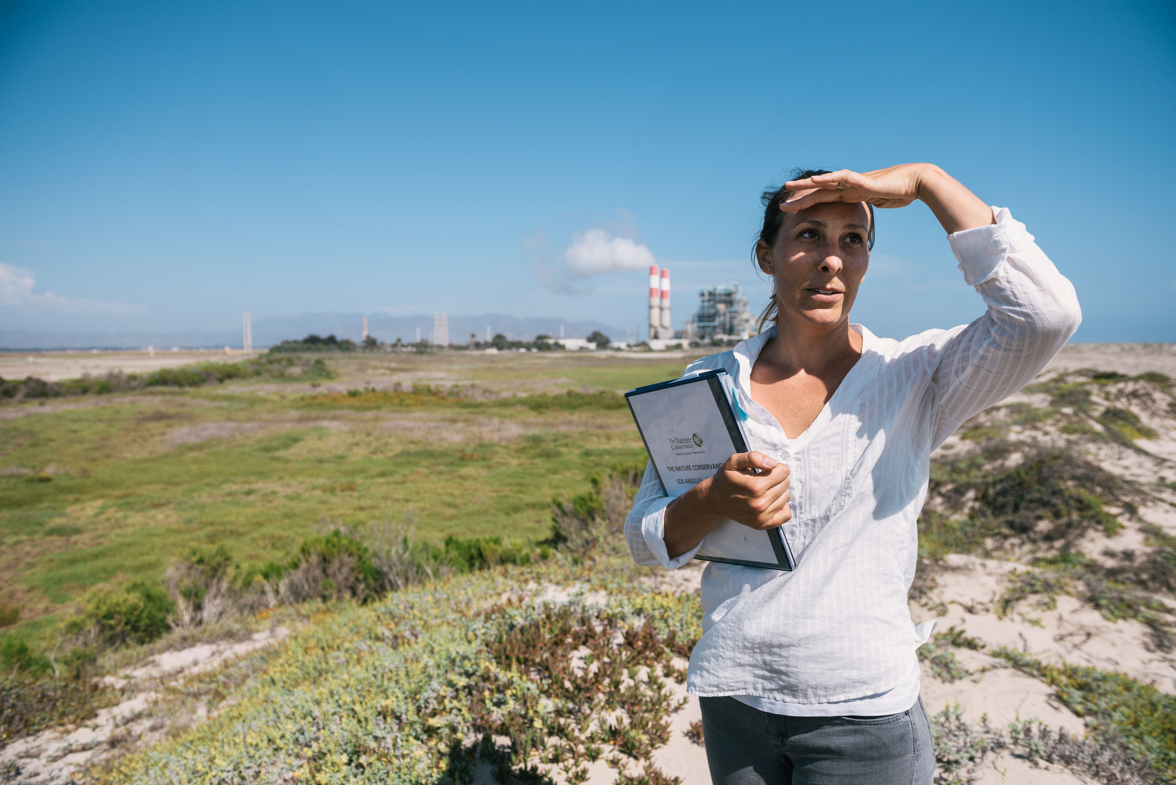 A woman holding a binder stands in a field and looks out in the distance while using her hand to shield her face from the sun.