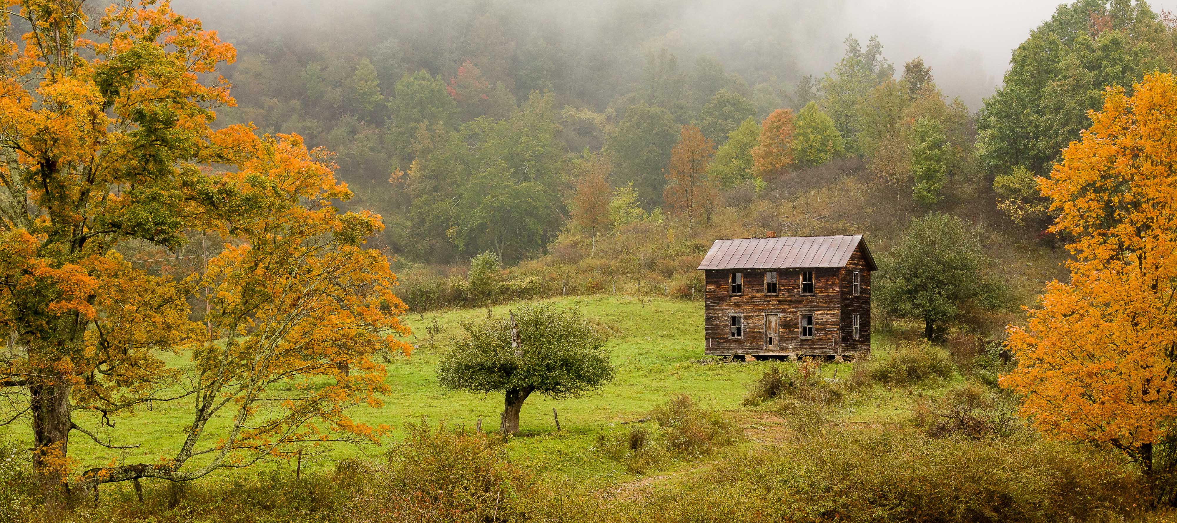 A dilapidated two story house with a tin roof sits alone in a small clearing surrounded by mature trees showing autumn colors.