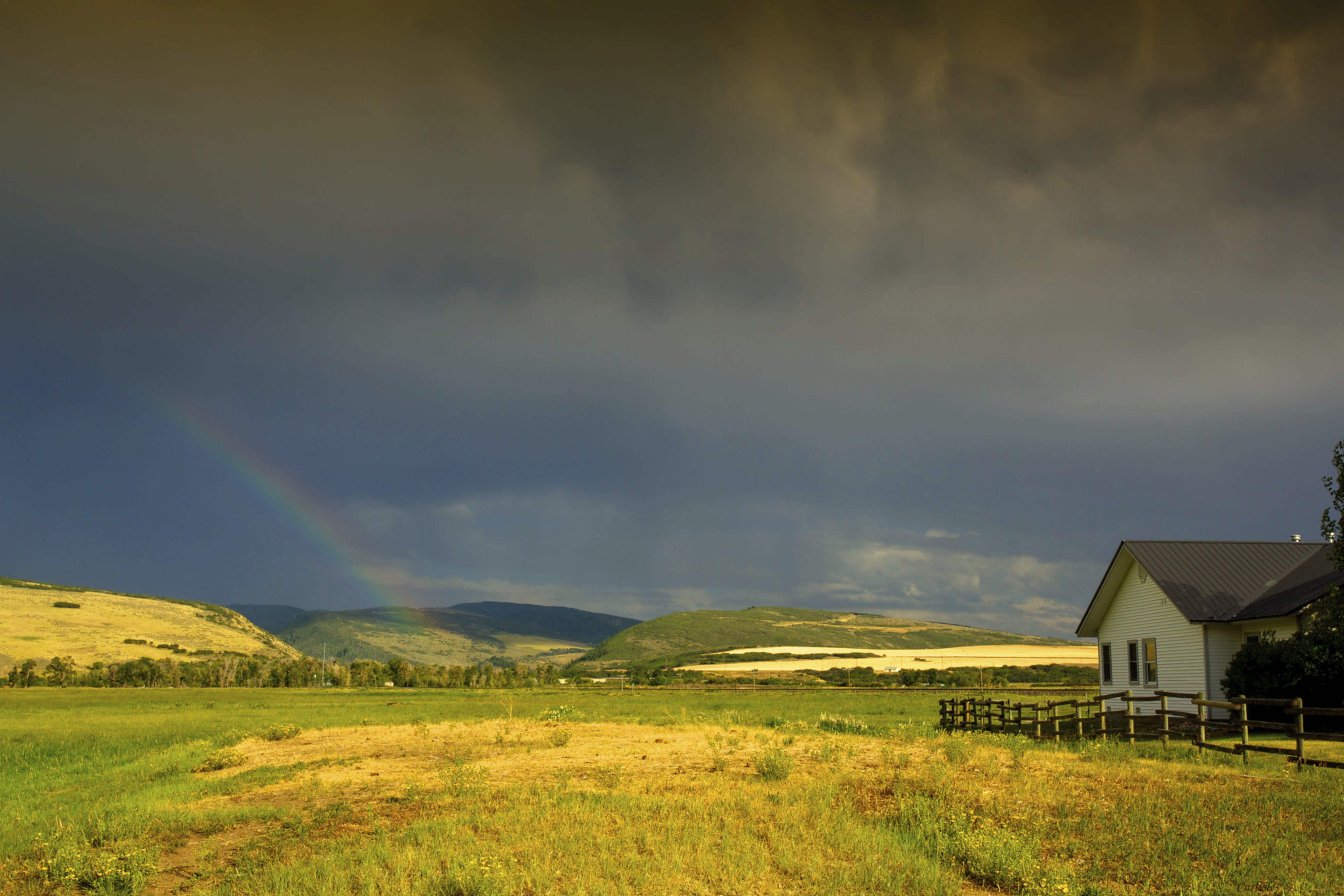 A house, wooden fence, fields and hills under a dark blue sky with a faint rainbow at Carpenter Ranch in Colorado.