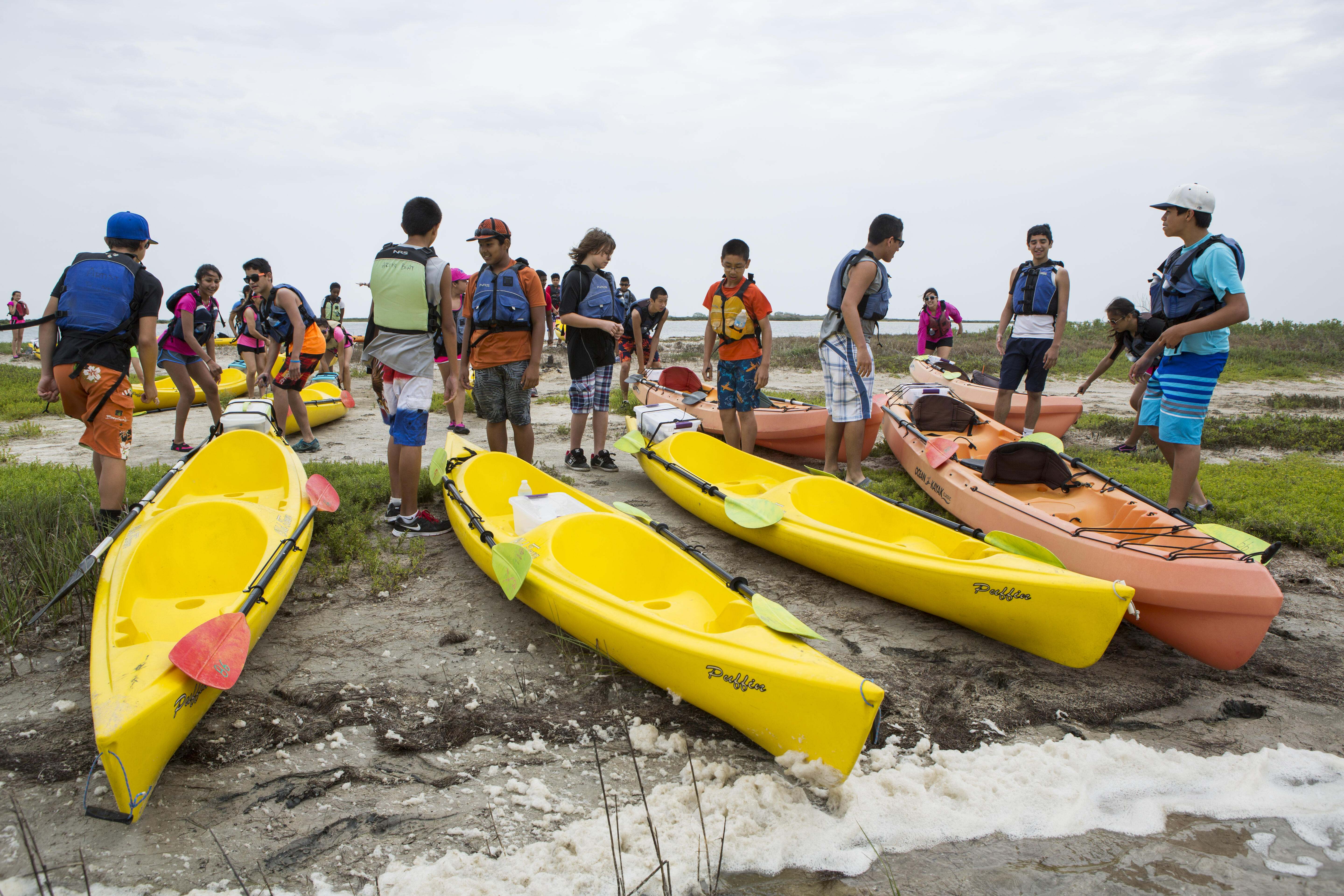 A large group of middle schoolers stands in life jackets on the beach, waiting to launch their yellow kayaks into the ocean. 