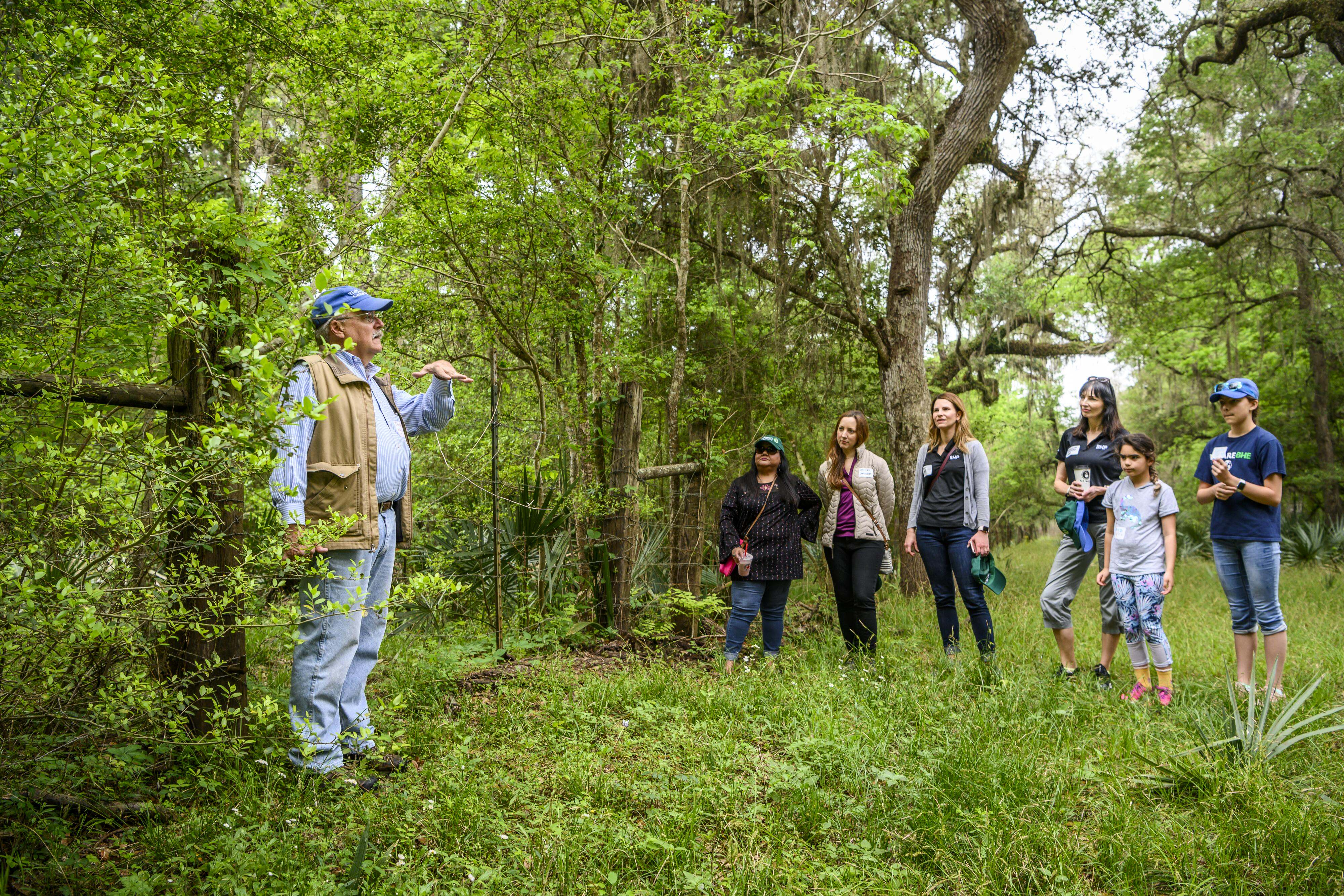 A man speaks to a group of 6 women in a green clearing surrounded by trees.