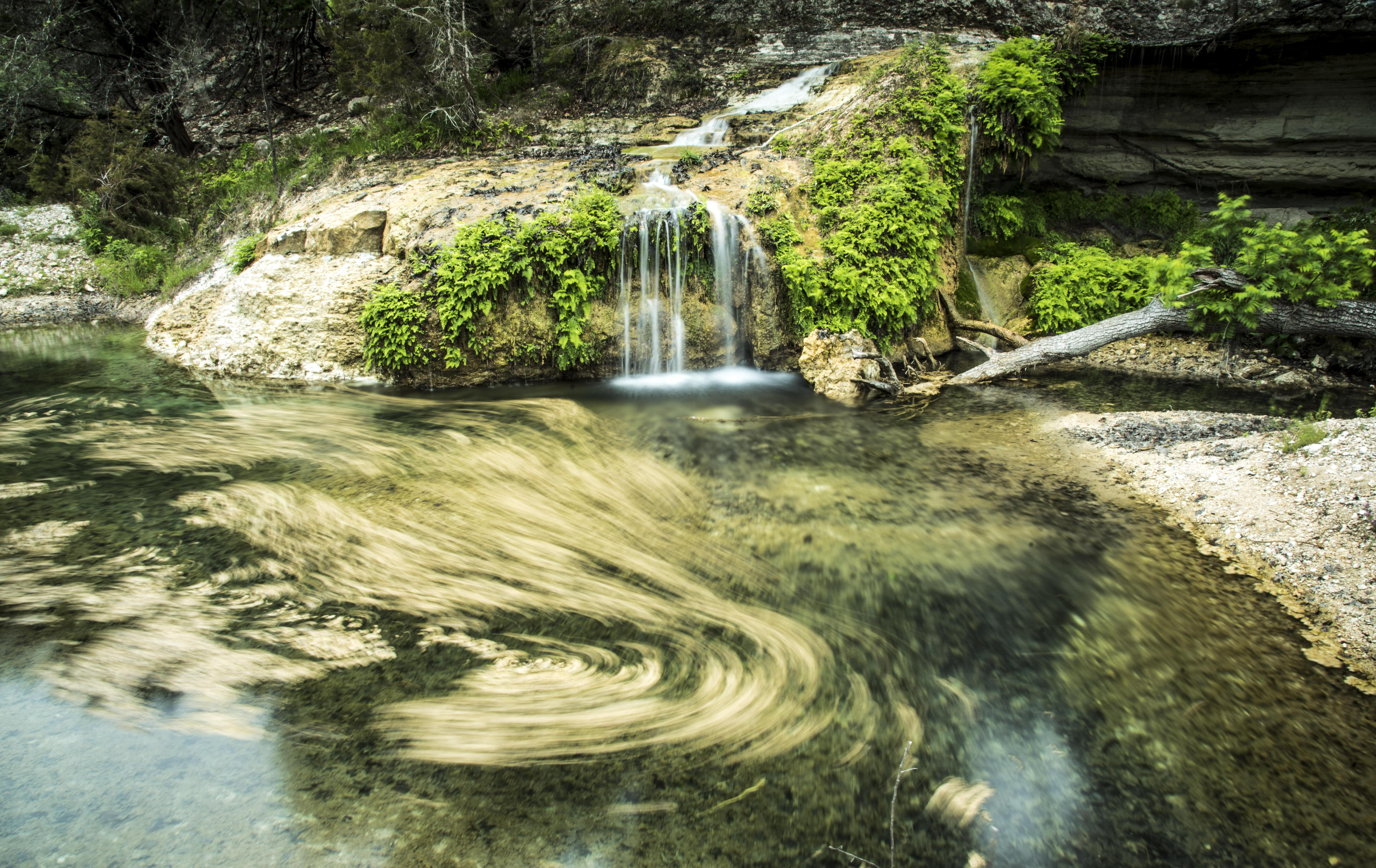 A stream trickles into a larger pool of clear, flowing freshwater, surrounded by bright green plants and moss on limestone outcroppings.