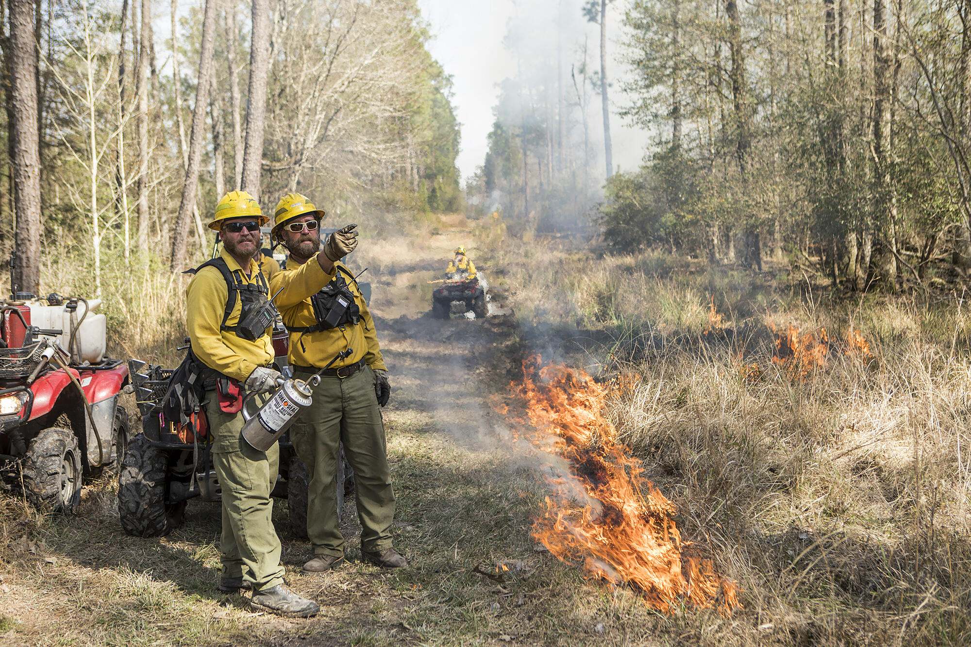 Two men in protective gear discuss burn plans as prescribed fire blazes at the edge of a firebreak. In the distance, a third fire practitioner rides on an ATV, surveying the burn.