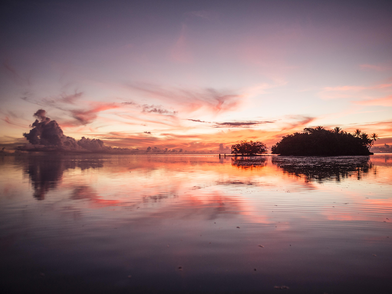 Landscape view of a large flat body of water with a small island; the sky and water are orange at sunset.