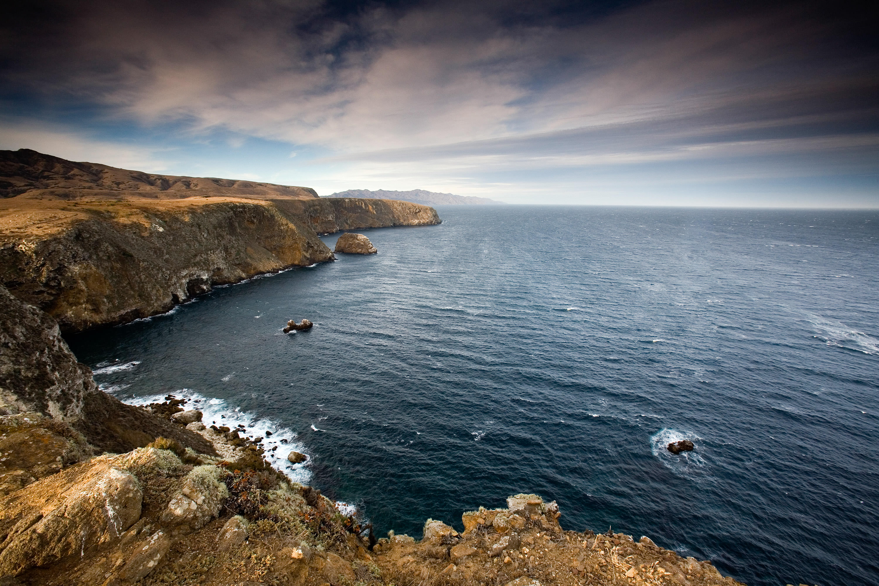 The blue water of the Pacific Ocean swells into waves that crash against tall coastal cliffs.