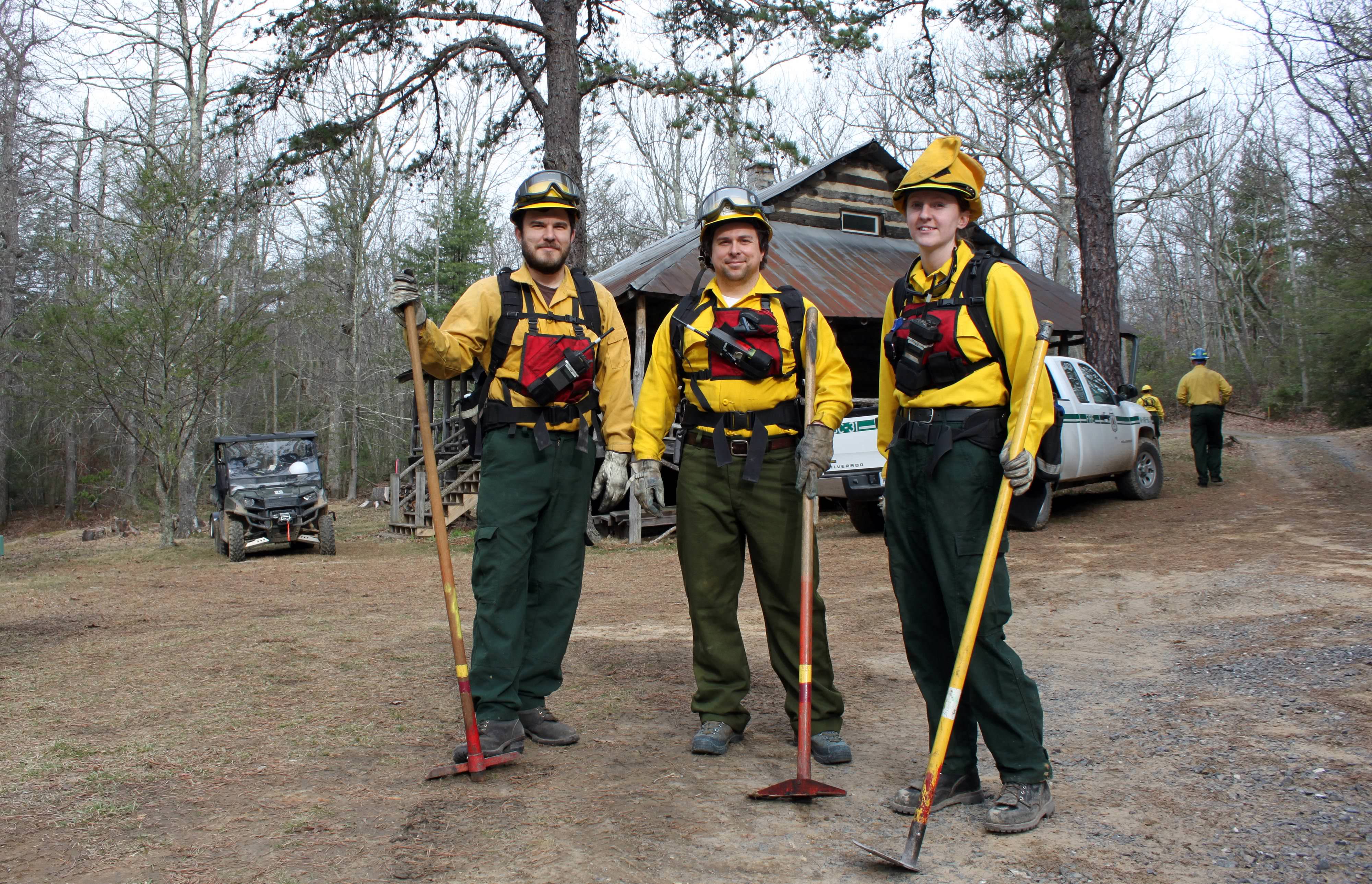 Three people pose together in front of a rustic cabin. They are wearing yellow fire-retardant gear and holding long-handled hoes. A white pickup truck is parked behind them.