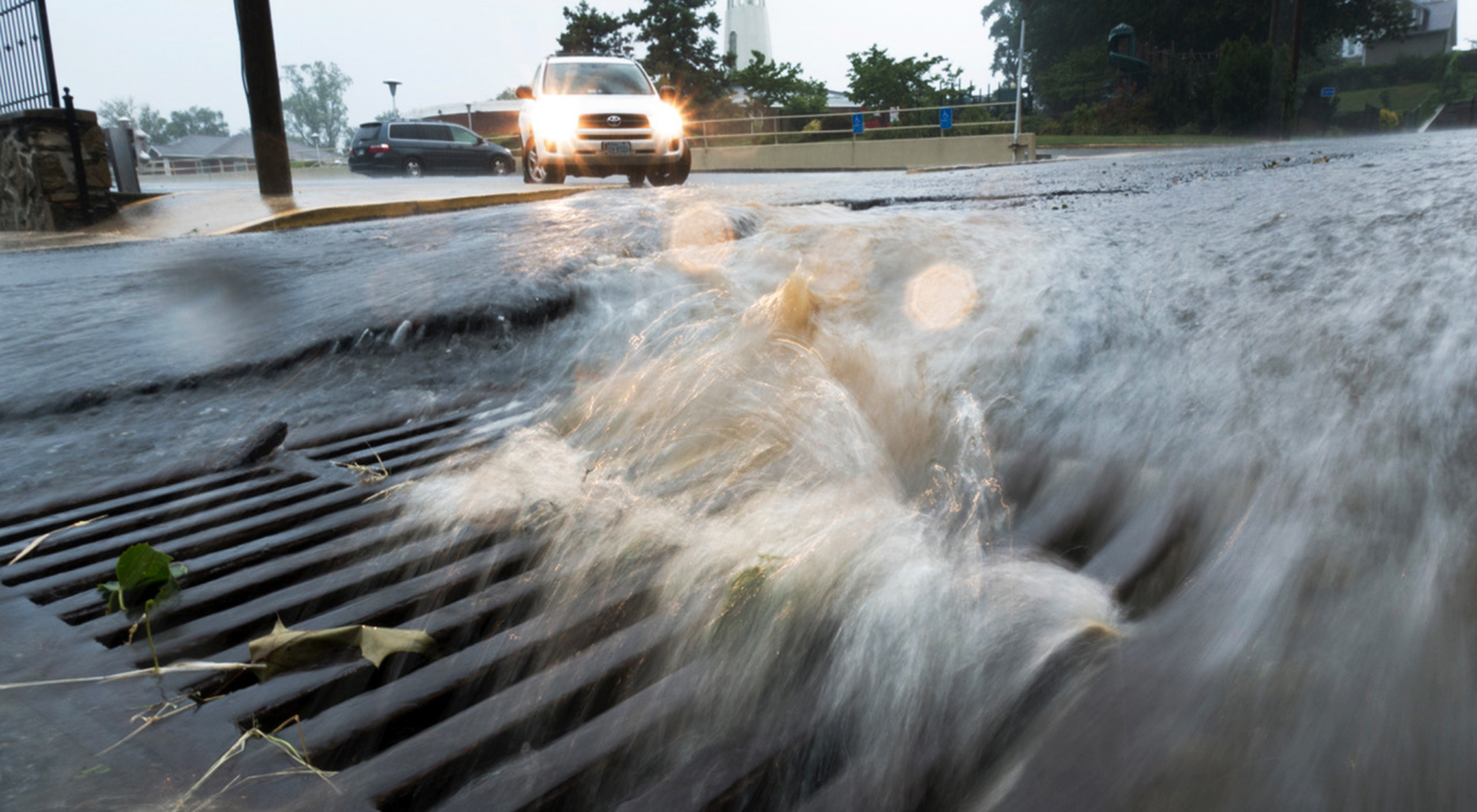 Storm water rushes down a city street into a storm drain. Cars in the background try to navigate the flooded street.