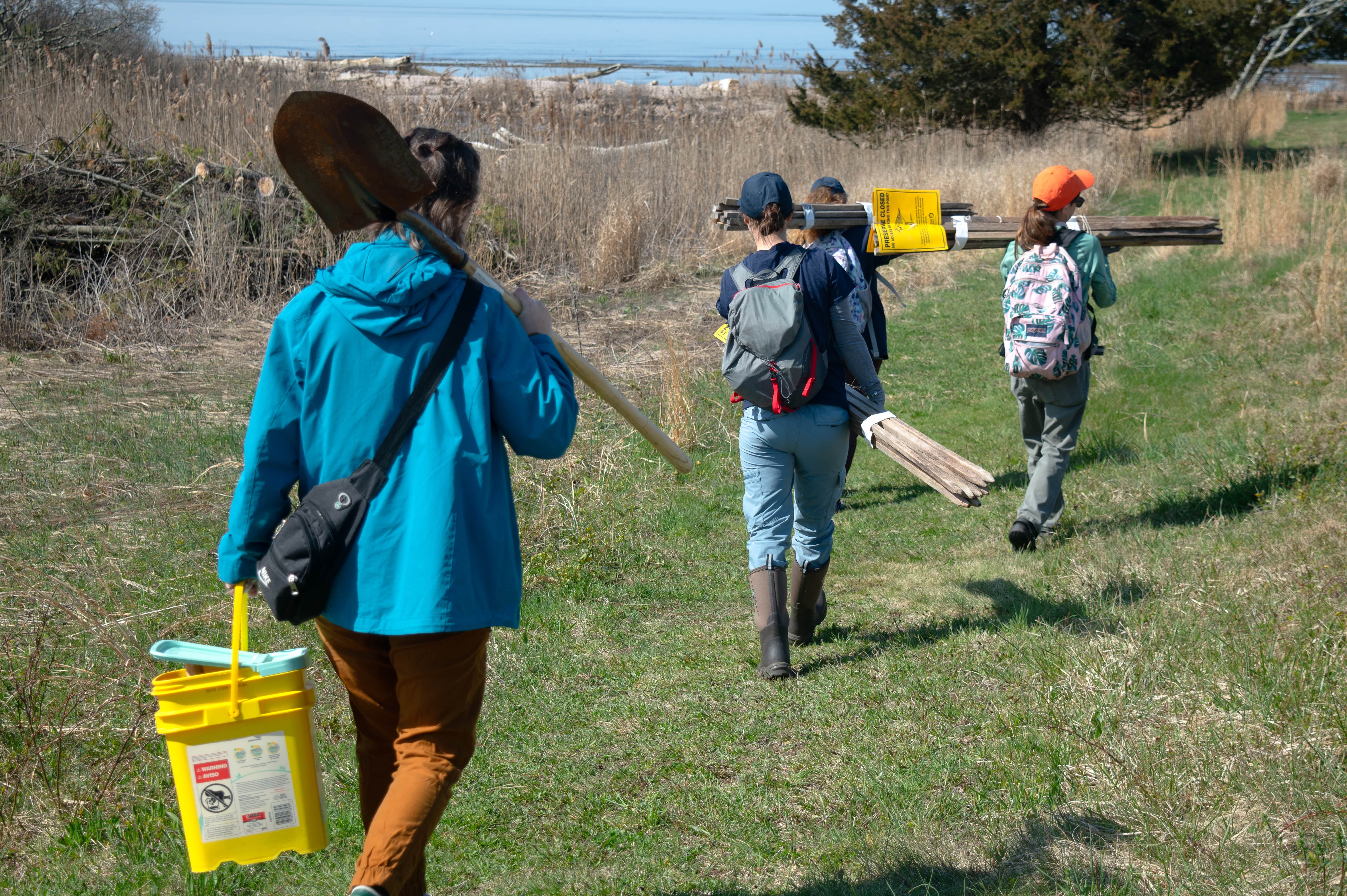 Several people holding equipment and tools walk down a grassy path.