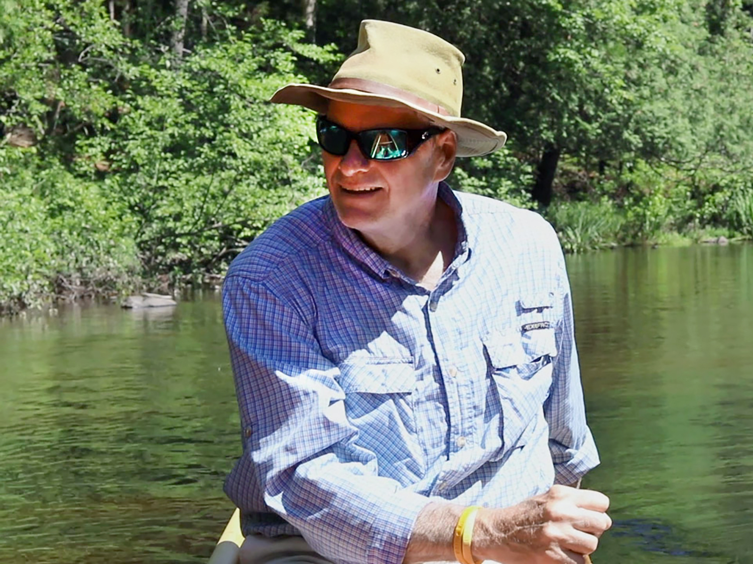Man in blue-checked shirt, sunhat and sunglasses paddles canoe on a river with forest in the background.