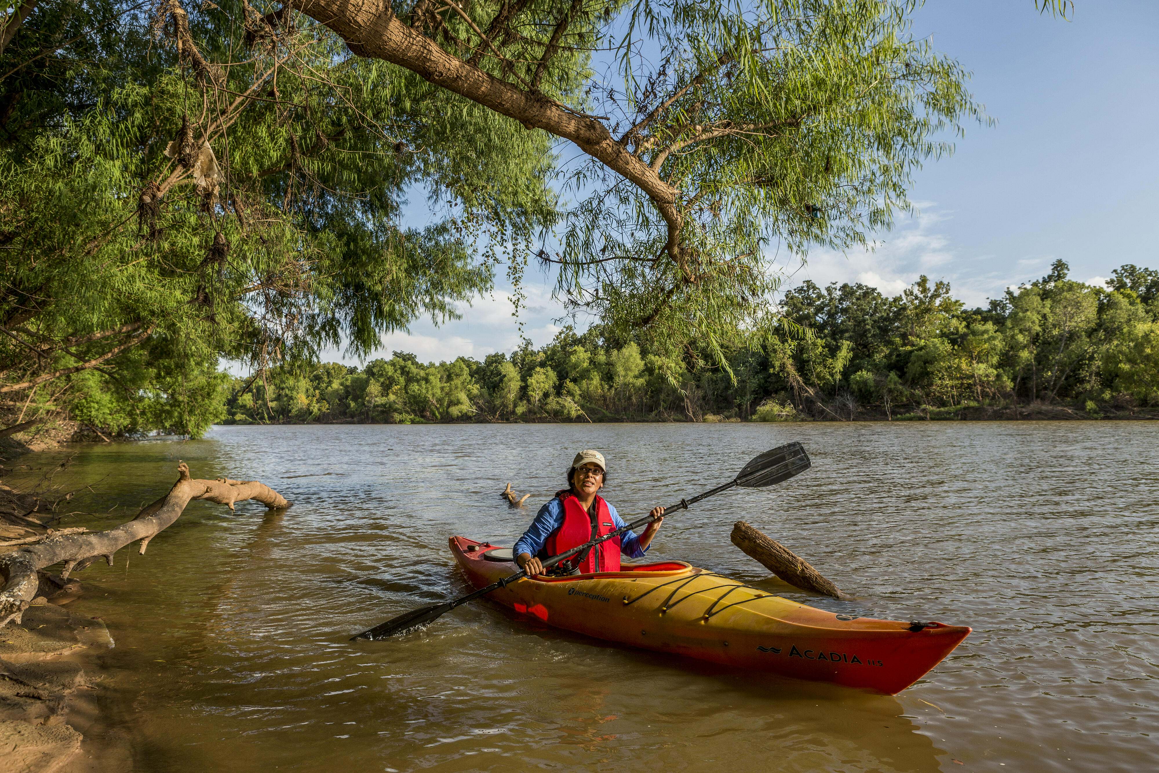 A woman in a hat paddles in a yellow kayak along a tree lined river.