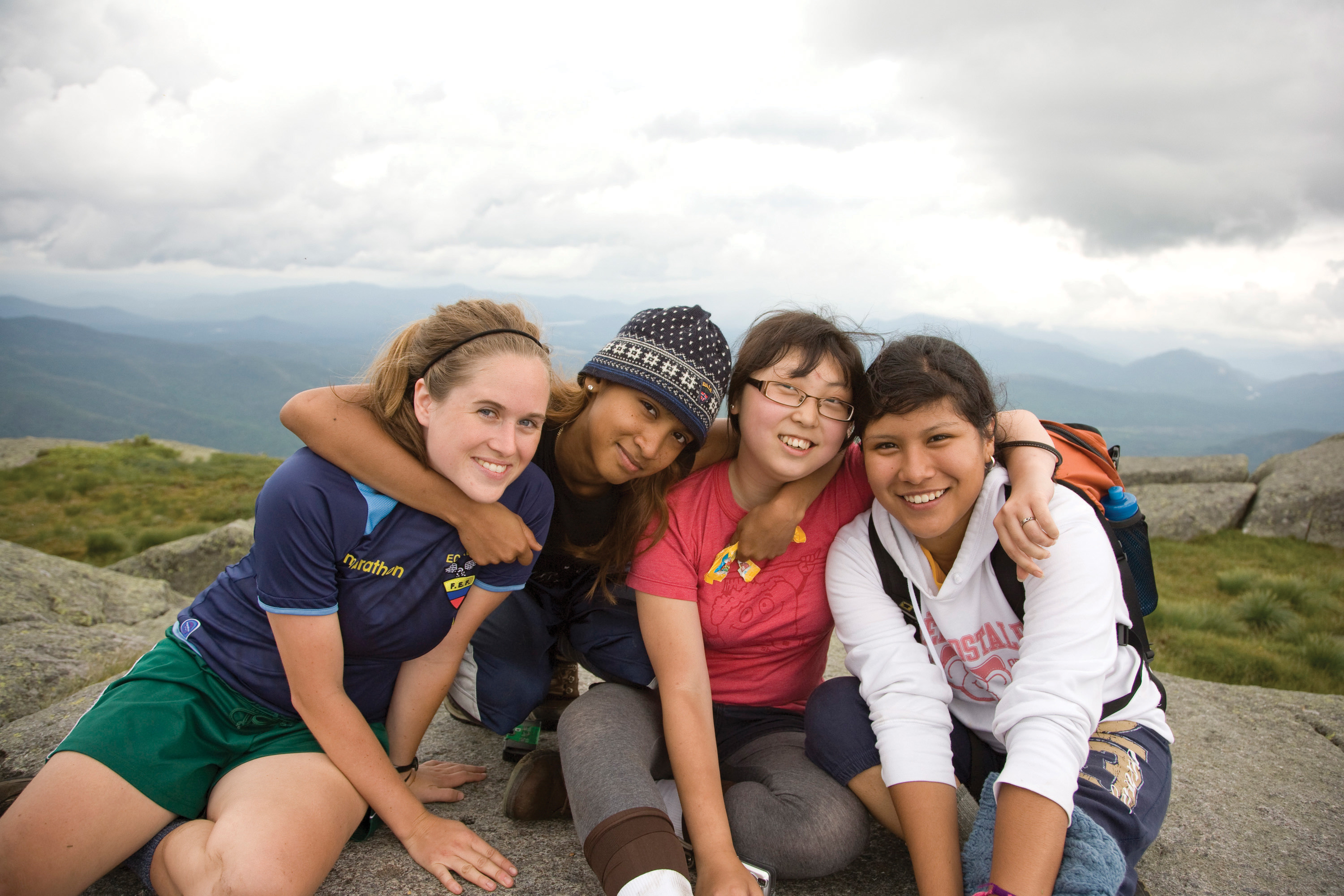 Four people sit together at a mountain overlook. There arms are linked around each others necks as they lean in and post together.